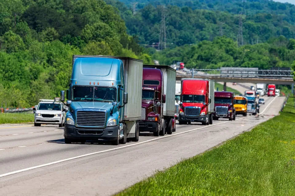 Line of multi-colored big rig trucks drivign down highway with other traffic including a yellow school bus.